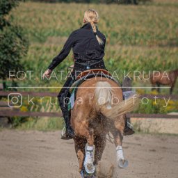 Kasia Krupa - Kobieta w czarnej kurtce i kapeluszu jeździ konno na piaszczystym terenie. Widać tylną część konia i kurz spod kopyt. Na zdjęciu widoczny znak wodny.