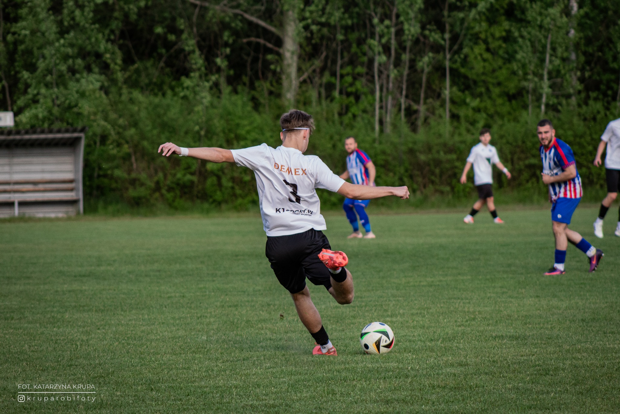 Dynamiczne ujęcie piłkarza w trakcie strzału na tle zielonego boiska i drzew. Widoczne logo sponsora na koszulce, akcja w trakcie meczu, inni zawodnicy w tle.