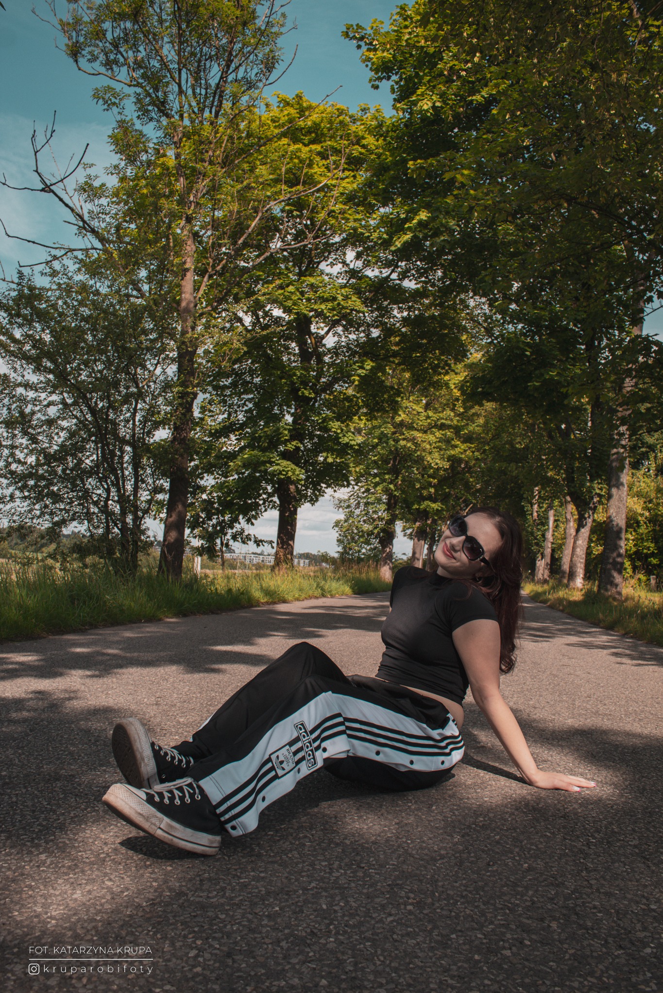 Portret kobiety w okularach przeciwsłonecznych, siedzącej na asfaltowej drodze otoczonej drzewami, w sportowym stroju Adidas i trampkach Converse, z podpisem autora.
