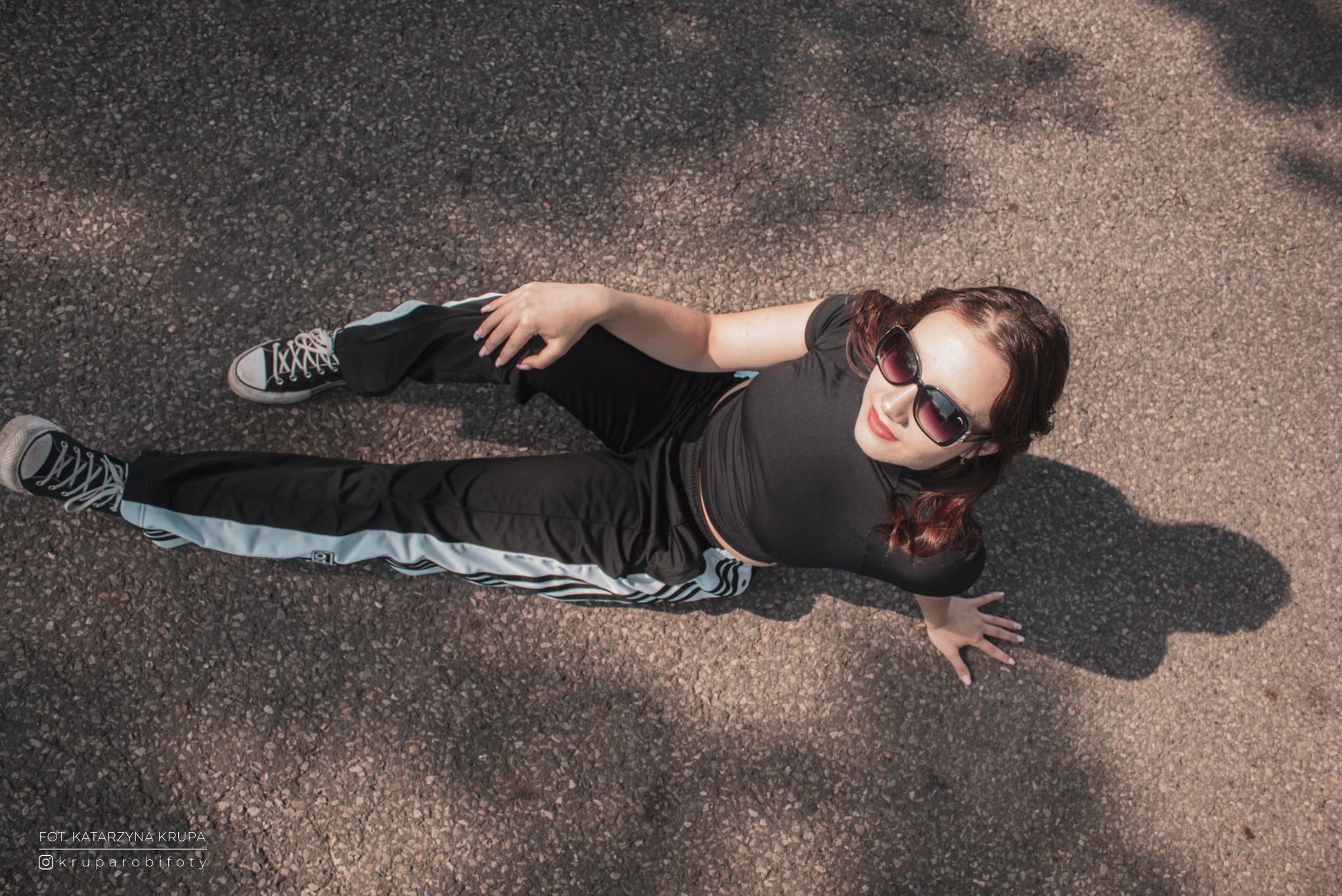 Zdjęcie z góry: młoda kobieta w czarnym stroju sportowym i okularach przeciwsłonecznych leży na szarym chodniku. Widoczne buty typu Converse i cień osoby.