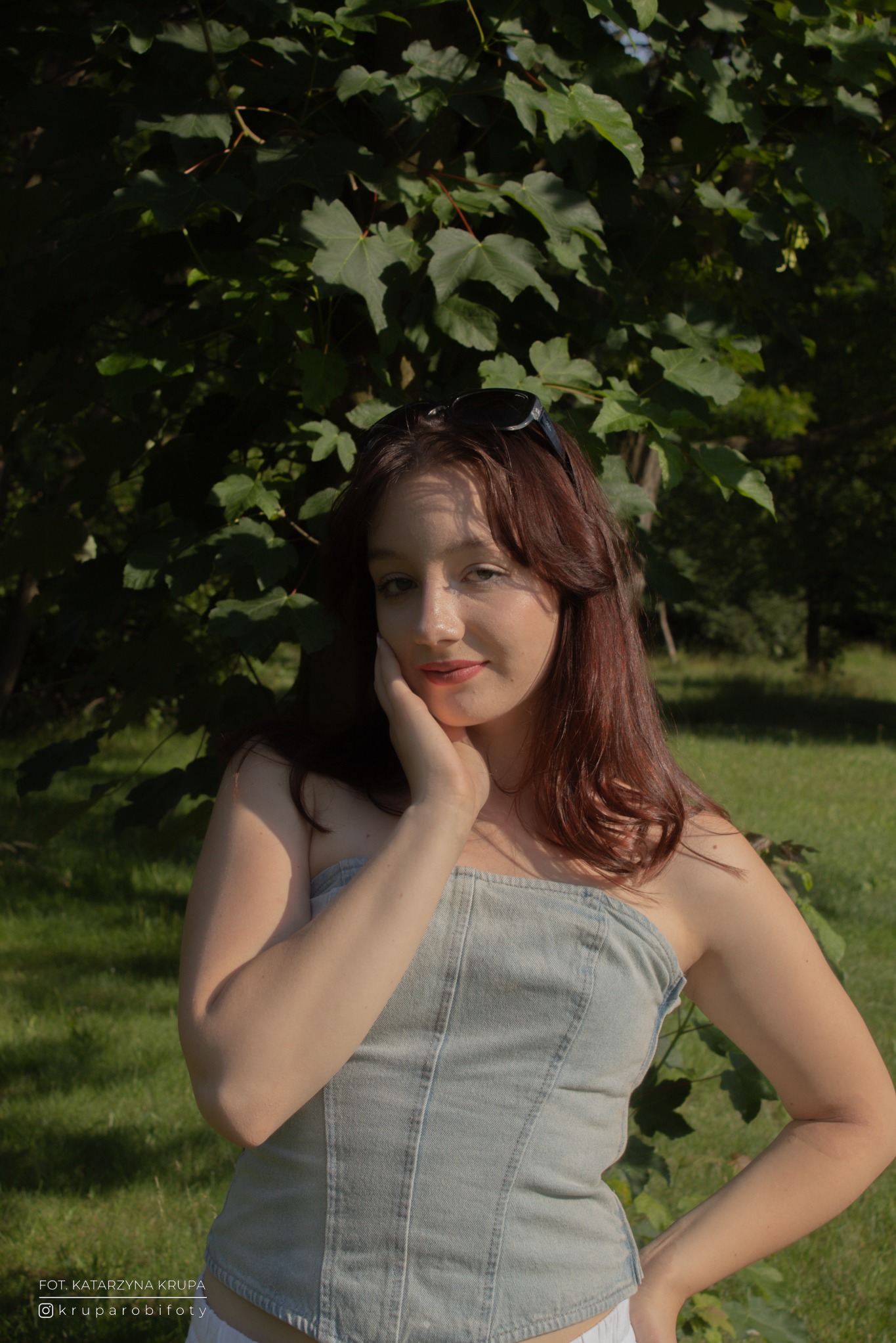 Portret kobiety w dżinsowym gorsecie na tle zieleni, z okularami na włosach. Delikatne światło podkreśla rysy twarzy, a naturalne tło dodaje swobody.