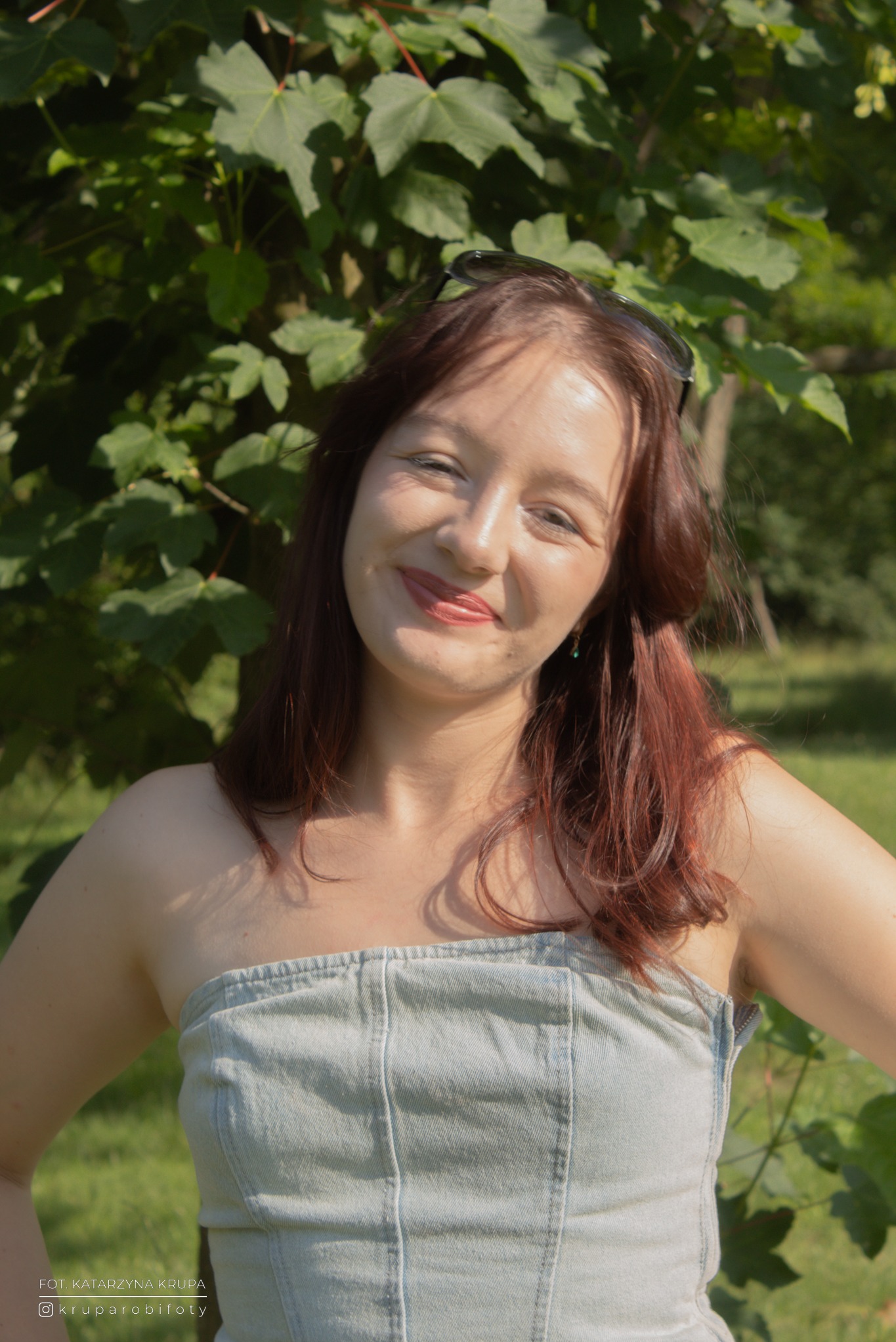 Portret uśmiechniętej kobiety w plenerze, z zielonym tłem liści, w letniej stylizacji. Okulary na głowie i delikatna biżuteria dodają uroku.