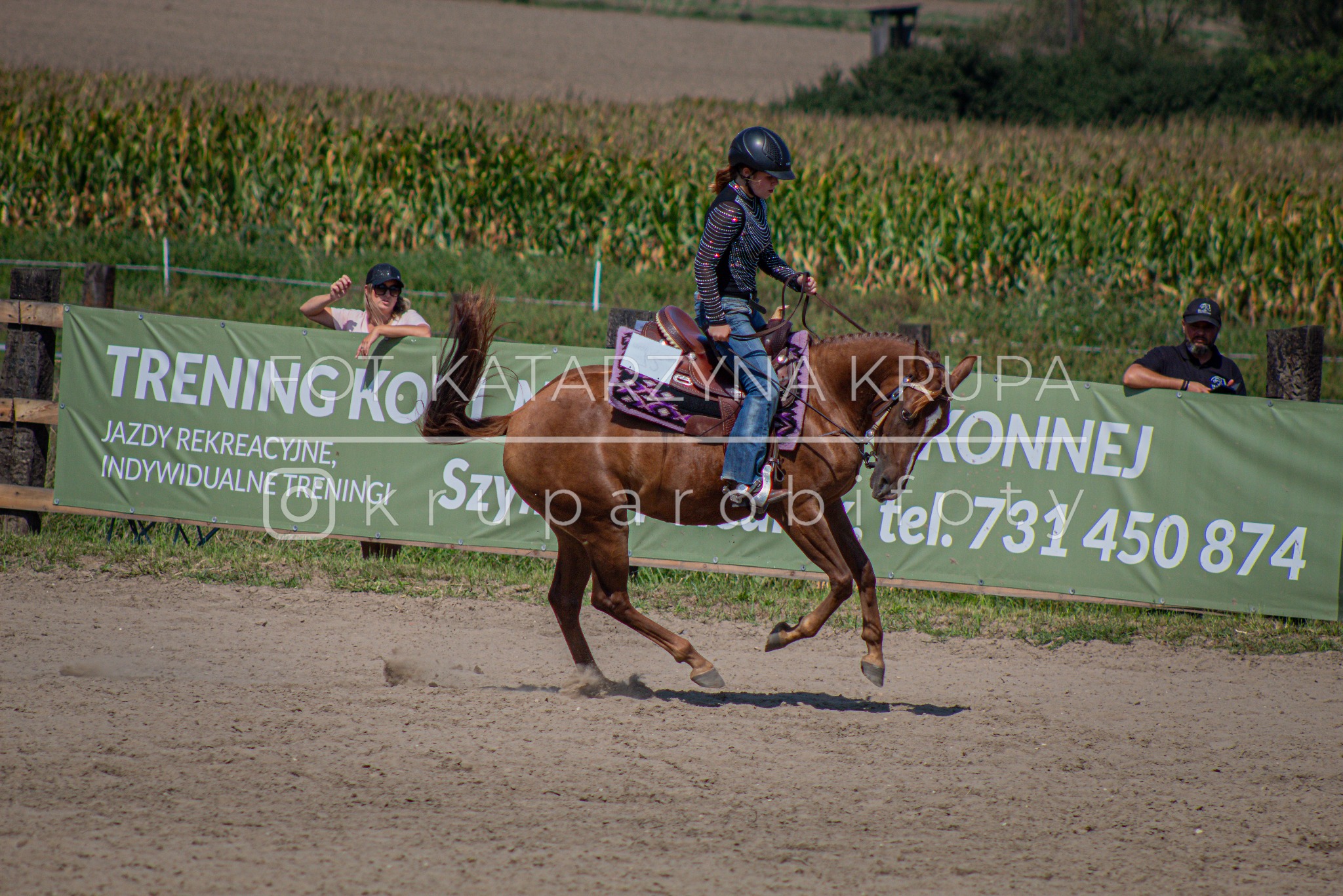 Kobieta w kasku jeździeckim na brązowym koniu w galopie na piaszczystym terenie, z widocznym banerem reklamowym i obserwatorami w tle.