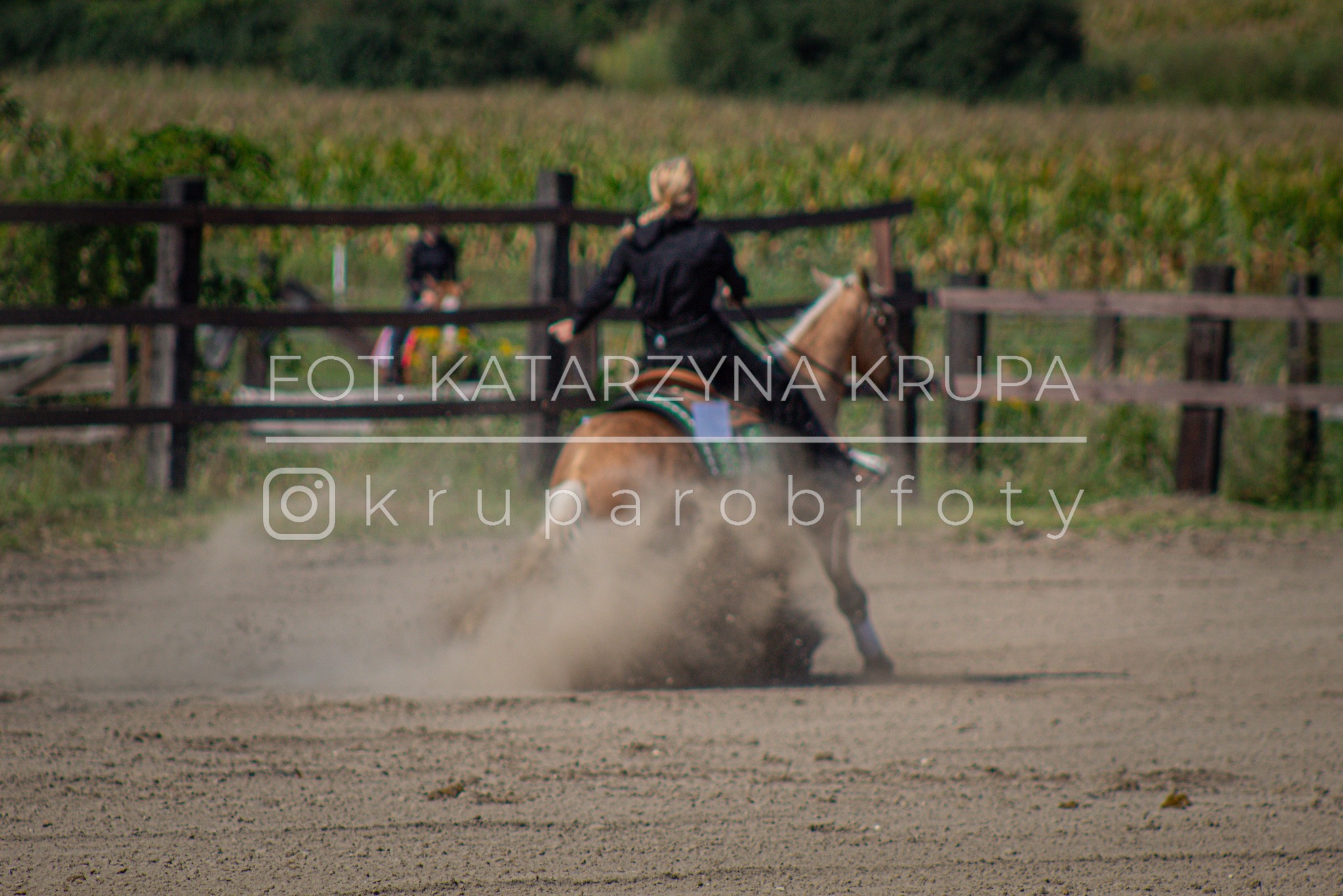 Dynamiczne ujęcie jeźdźca na koniu, unoszący się pył, w tle drewniane ogrodzenie i pole. Fotografia z logo fotografa Katarzyny Krupy.