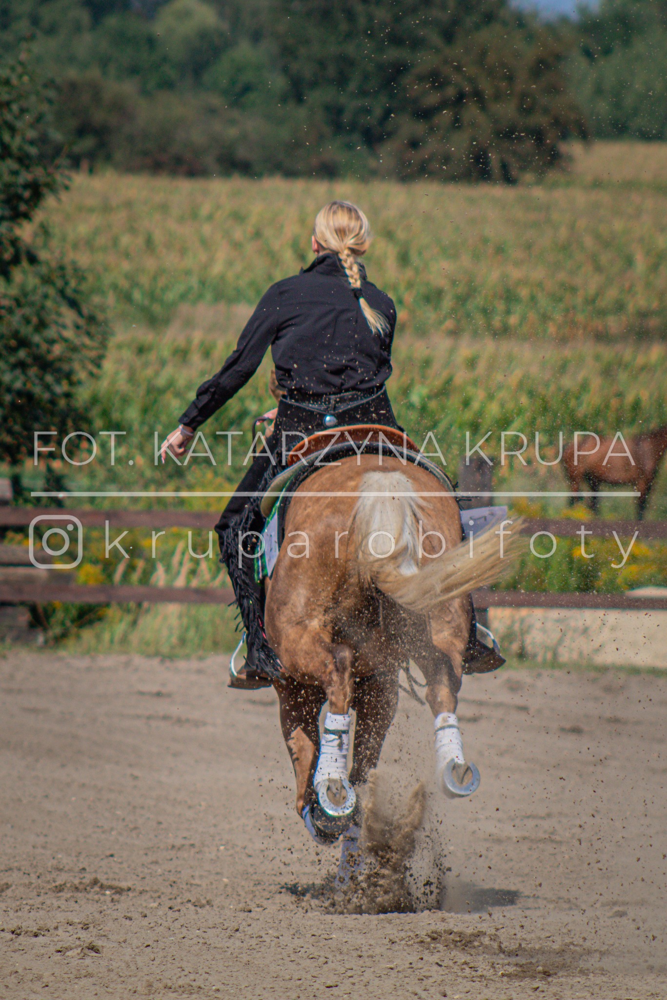 Kobieta w czarnej kurtce i kapeluszu jeździ konno na piaszczystym terenie. Widać tylną część konia i kurz spod kopyt. Na zdjęciu widoczny znak wodny.