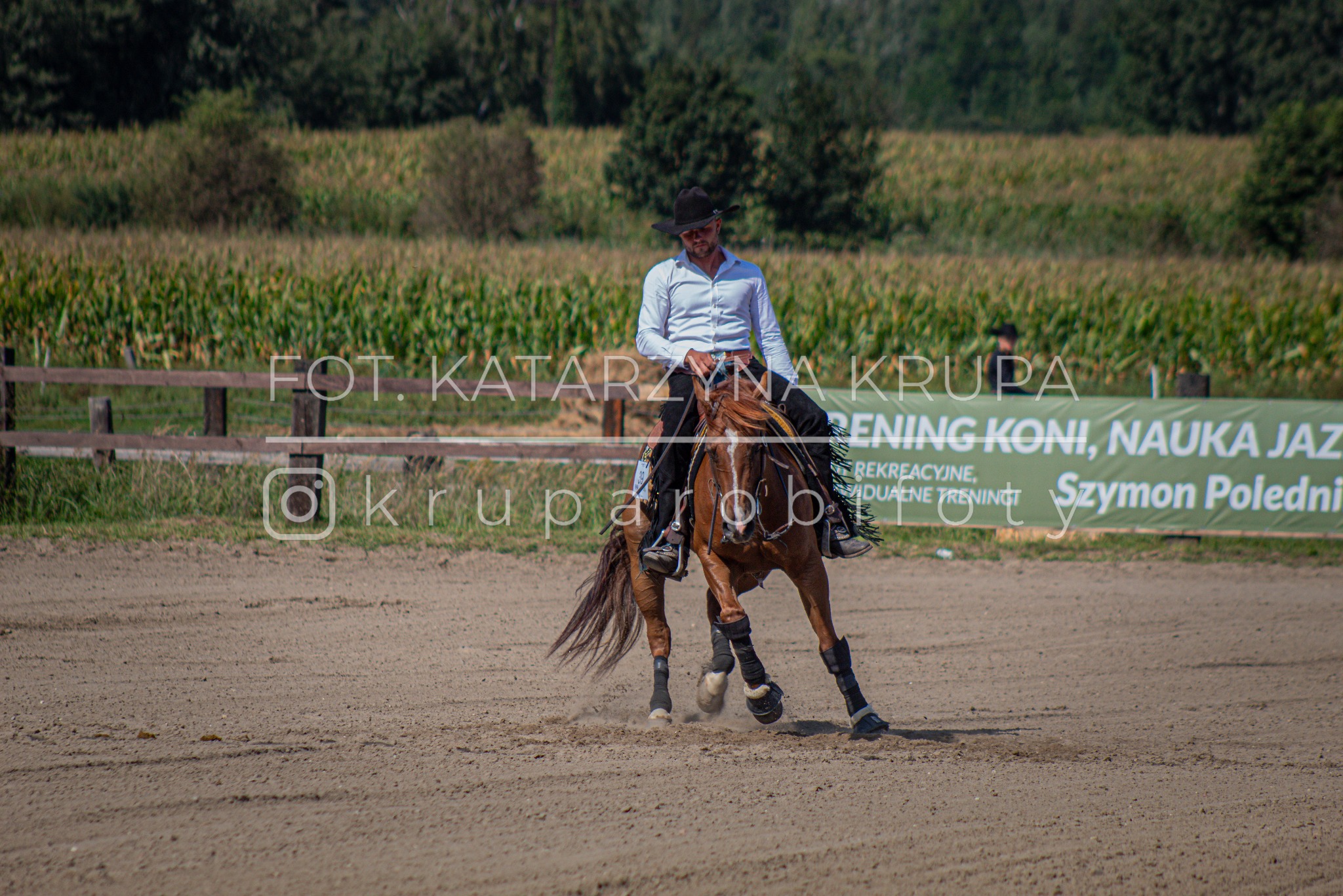 Mężczyzna w kapeluszu i białej koszuli jeździ na brązowym koniu na piaszczystym terenie; w tle pole kukurydzy i baner z napisem 'Trening koni, nauka jazdy'.