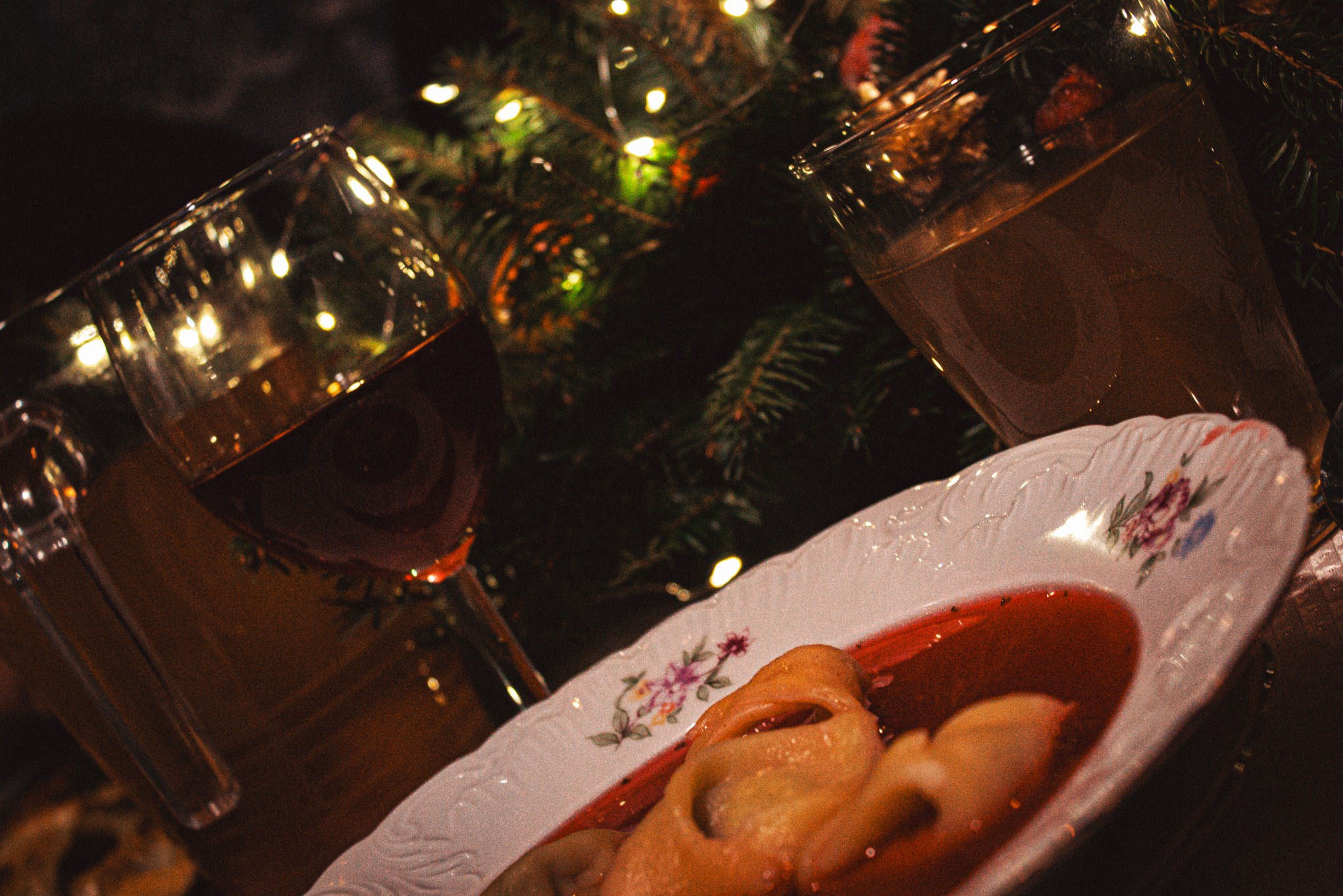 Świąteczny stół: barszcz z uszkami na zdobionym talerzu, obok kieliszek czerwonego wina i szklanka z kompotem, w tle choinka z lampkami, ciepłe, nastrojowe światło.