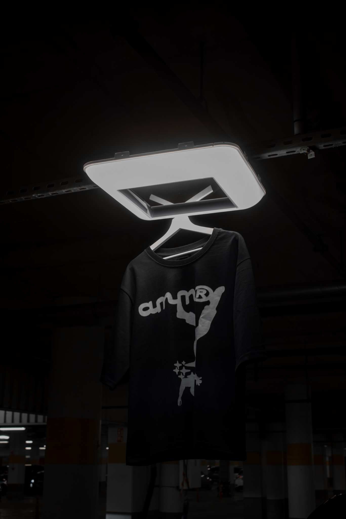 Czarna koszulka z białym nadrukiem marki 'ammr' w nietypowym ujęciu, zawieszona pod lampą na tle parkingu podziemnego, tworzy minimalistyczny, miejski klimat.