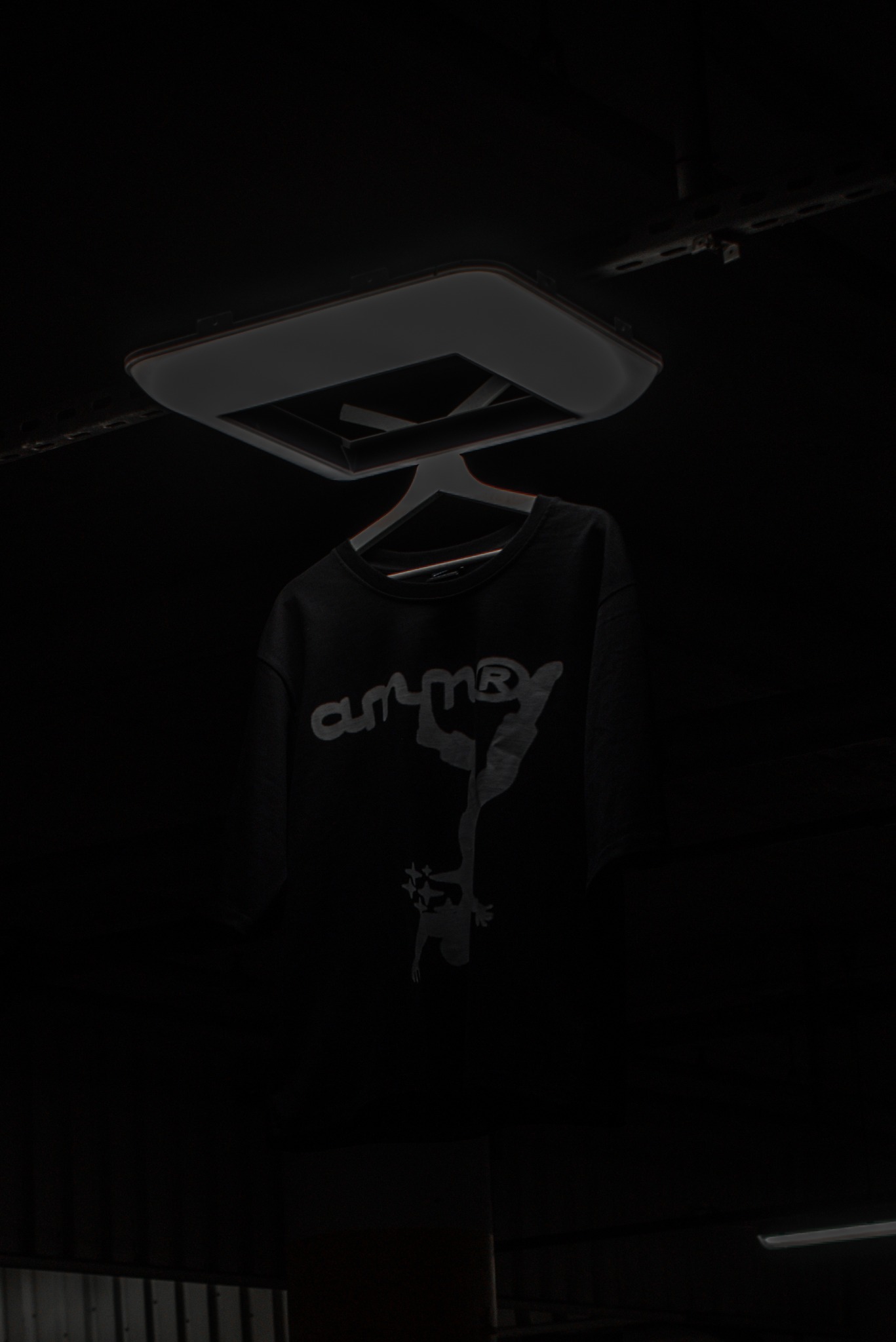 Czarna bluza z szarym nadrukiem na wieszaku, oświetlona od góry, na ciemnym tle. Ujęcie z dołu, widoczny fragment sufitu.