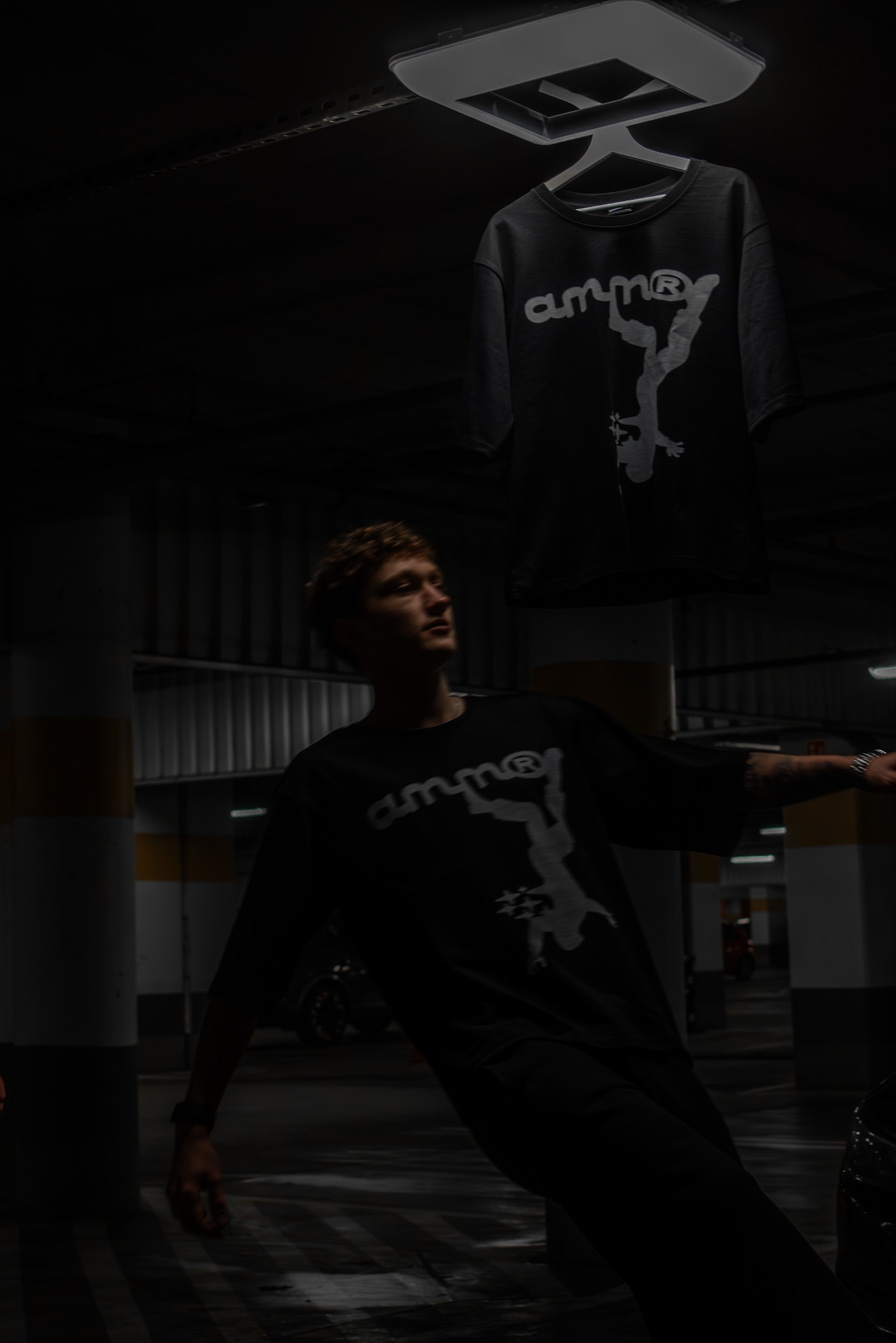 Mężczyzna w czarnej koszulce z nadrukiem, pozujący w garażu podziemnym, z wiszącą koszulką z tym samym wzorem. Nietypowe oświetlenie i kąt ujęcia.