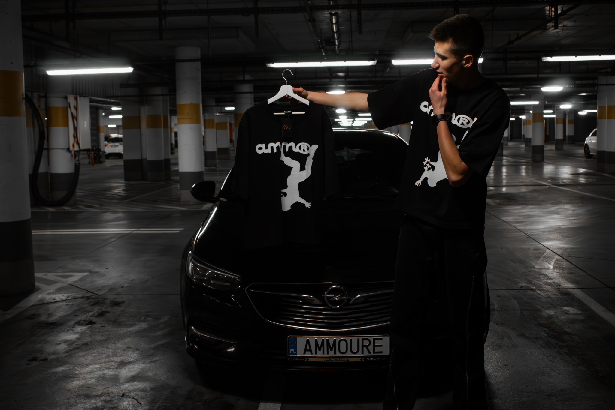 Mężczyzna prezentuje czarną koszulkę z białym nadrukiem na wieszaku, stojąc na tle czarnego samochodu na parkingu podziemnym. Ubrany w pasującą koszulkę z tym samym wzorem.