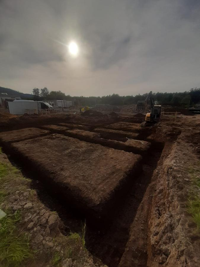 Wykopane fundamenty pod budowę domu jednorodzinnego, z widocznym sprzętem budowlanym w tle i zachmurzonym niebem. Etap prac ziemnych.