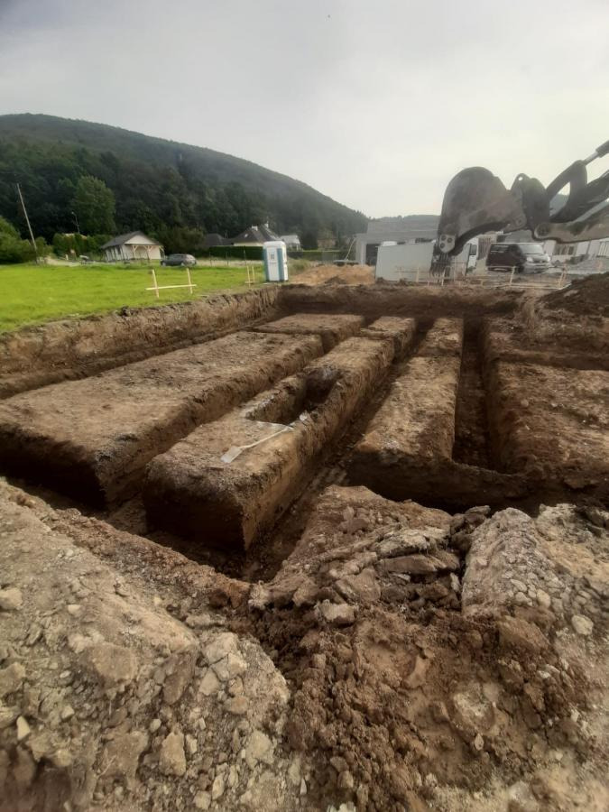 Wykopane rowy pod fundamenty na tle zieleni i budynku. Widoczny sprzęt budowlany i tymczasowa toaleta. Ujęcie z bliska na wykopany teren.
