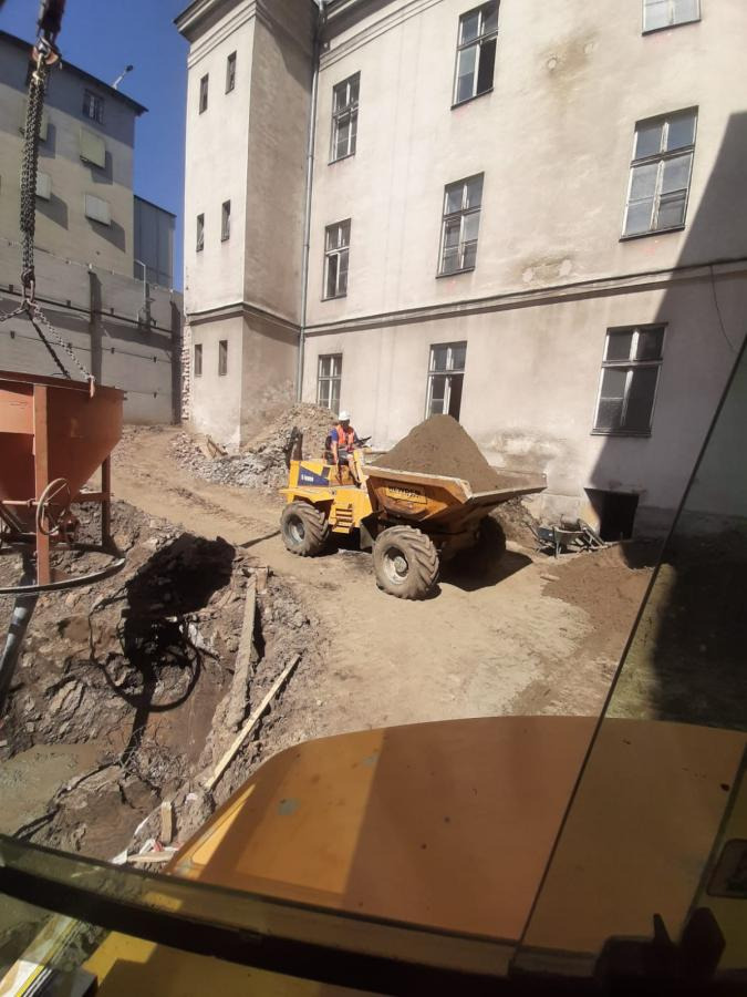 Żółta wywrotka przewozi ziemię na terenie budowy przy starym budynku. W tle widoczny dźwig i wykop. Operator w kasku na maszynie.