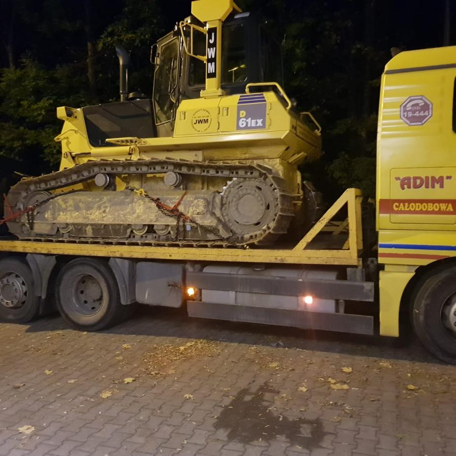 Transport żółtego spychacza JWM D61EX na niskopodwoziowej platformie ciężarówki ADIM, widok z boku, nocne oświetlenie, teren z kostki brukowej.
