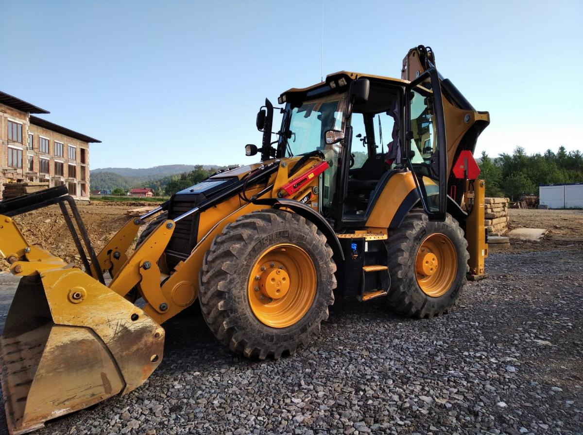 Żółto-czarna koparko-ładowarka na żwirowym terenie budowy, z budynkiem w tle i otwartymi drzwiami kabiny, w słoneczny dzień.