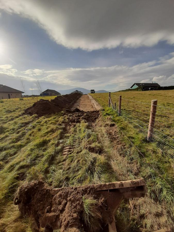 Wąski wykop w ziemi na nierównym terenie zielonym, z widocznym fragmentem maszyny wykopowej w pierwszym planie i zabudowaniami w tle, w słoneczny dzień.