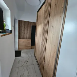 Nowoczesna szafa wnękowa z drewnianymi lamelami w przedpokoju z marmurową podłogą, oświetlona okrągłą lampą sufitową.