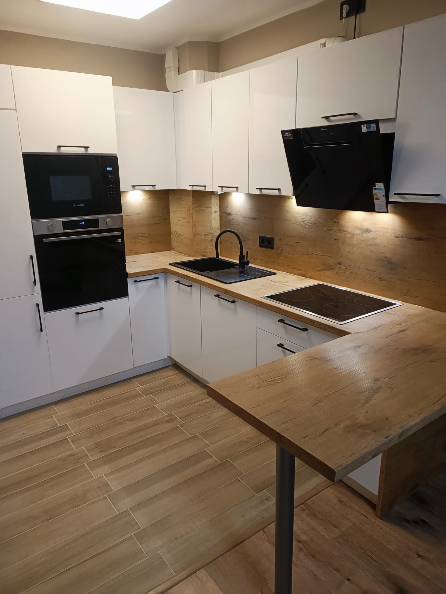 Nowoczesna kuchnia z białymi szafkami, drewnianym blatem i czarnymi akcentami, w tym zlew i okap. Na podłodze płytki imitujące drewno.