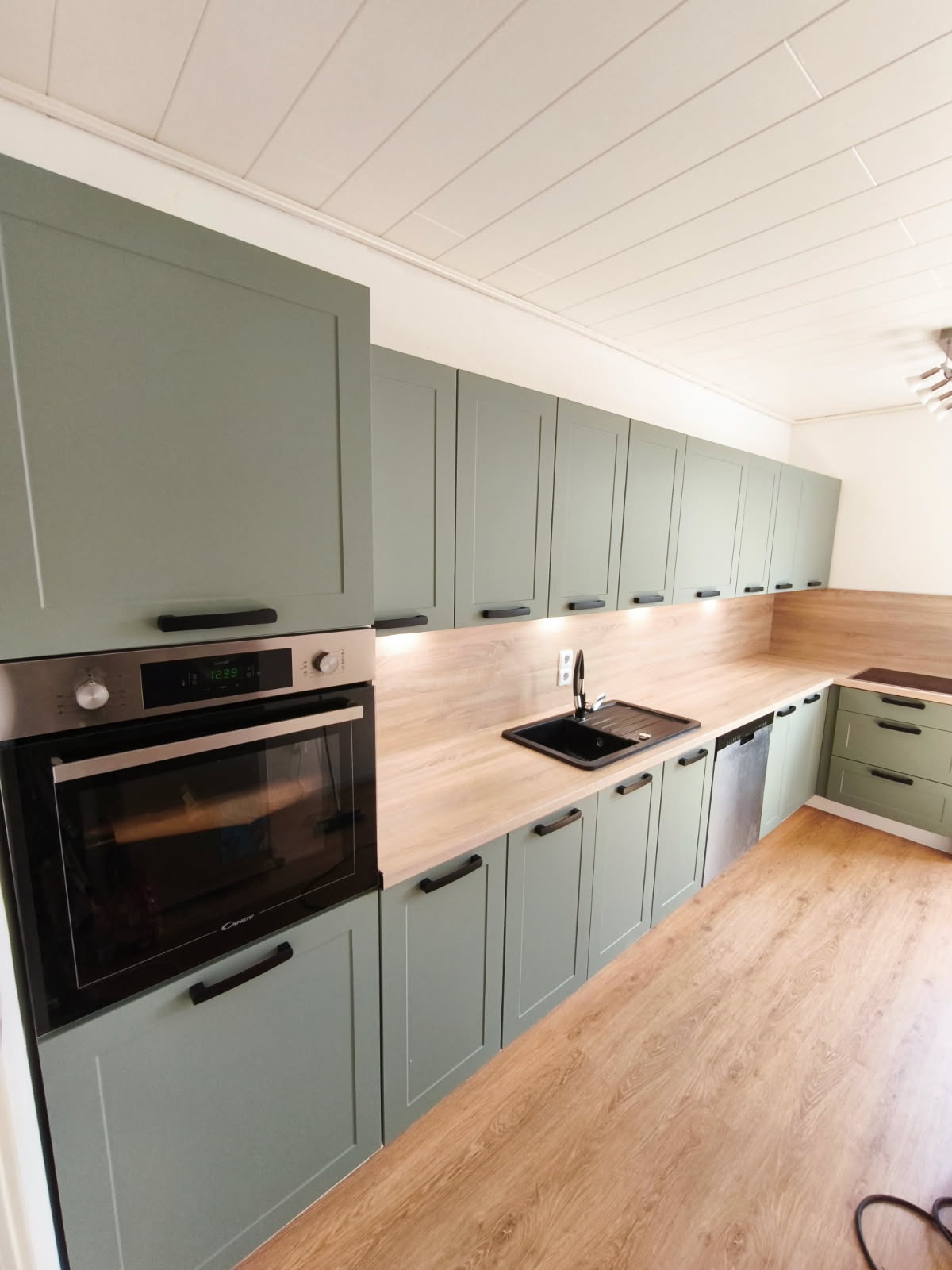 Nowoczesna kuchnia w odcieniach zieleni z drewnianym blatem i czarnym zlewem. Widoczny piekarnik Candy i oświetlenie podszafkowe. Minimalistyczny design, jasne wnętrze.