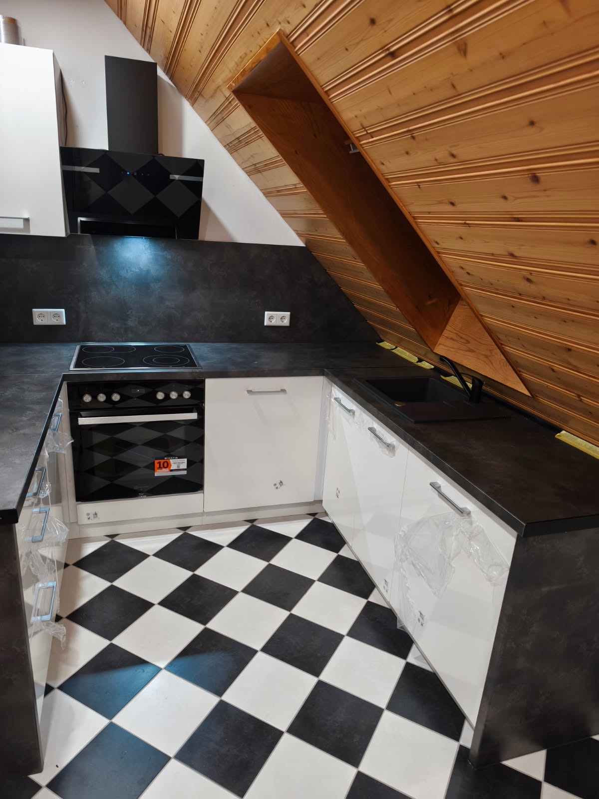 Kuchnia z białymi szafkami, czarnym blatem i płytkami w szachownicę, umieszczona na poddaszu z drewnianą boazerią. Widoczny piekarnik z płytą grzewczą i okap.