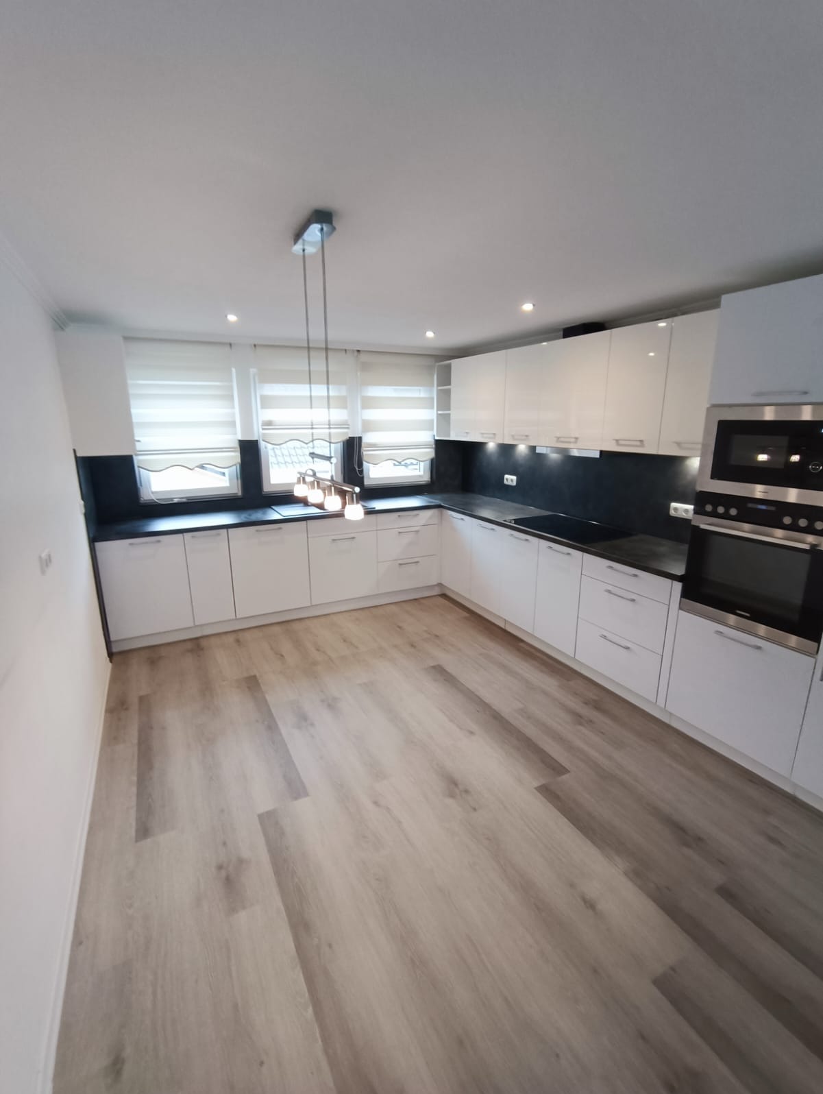 Jasna kuchnia w kształcie litery U z białymi, lakierowanymi frontami, czarnym blatem i drewnianą podłogą. Nad blatem wiszą trzy lampy z przezroczystymi kloszami.