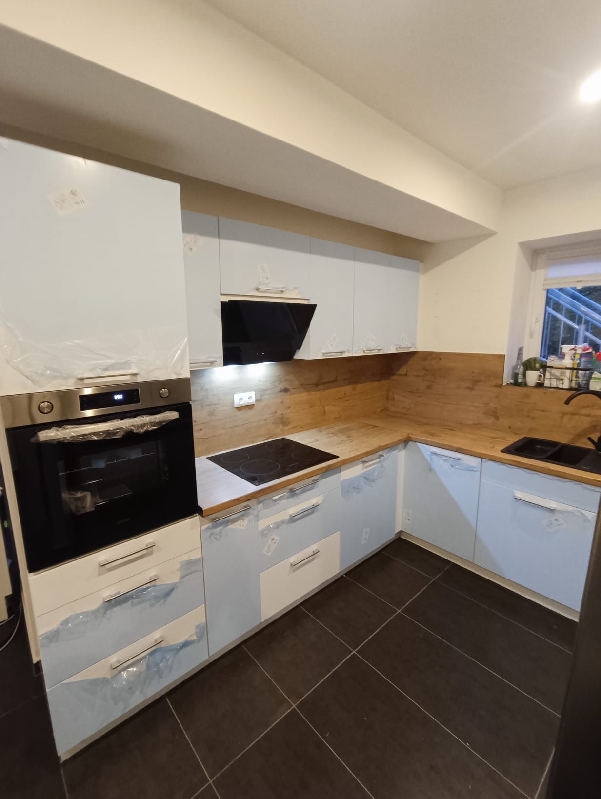 Jasna, narożna kuchnia z drewnianym blatem i czarną płytą grzewczą. Fronty szafek zabezpieczone folią ochronną. Widoczny piekarnik i okap.