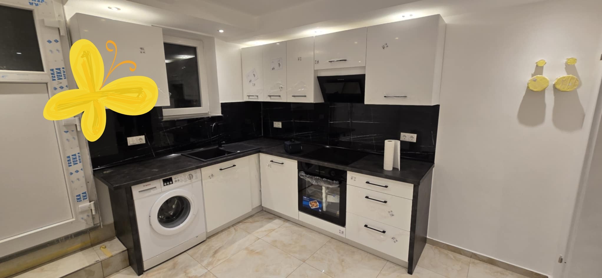 Nowoczesna kuchnia z białymi, lakierowanymi frontami, czarnym blatem i marmurowym backsplashiem. Pralka w zabudowie obok zlewu. Minimalistyczny design, subtelne oświetlenie.