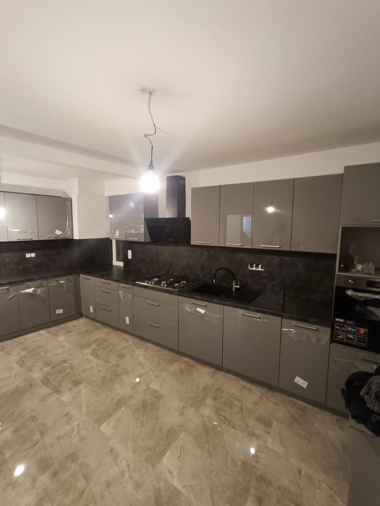 Szara kuchnia na wymiar z czarnym blatem i płytą gazową, fronty częściowo zabezpieczone folią, marmurowa podłoga, widok na całe pomieszczenie.