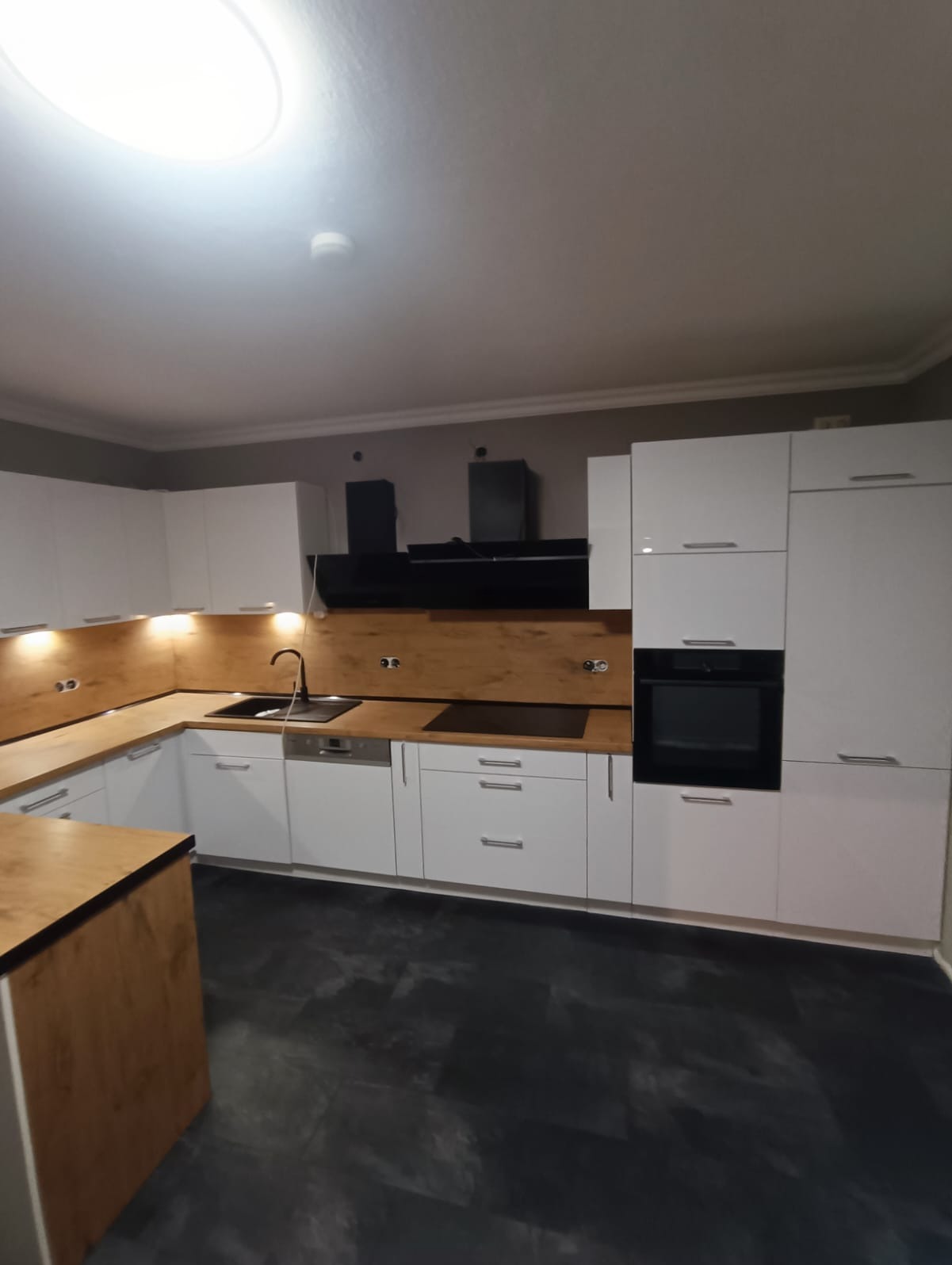 Nowoczesna biała kuchnia z drewnianym blatem i czarnymi akcentami, w tym okapem i piekarnikiem, na tle ciemnej podłogi.