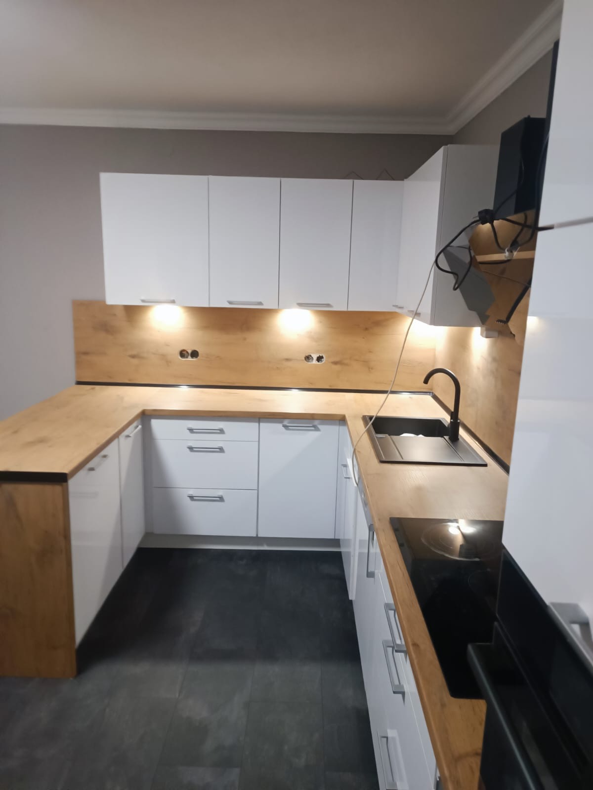 Nowoczesna kuchnia w kształcie litery U z białymi szafkami i drewnianym blatem. Oświetlenie podszafkowe. Czarny zlew i płyta indukcyjna. Ciemna podłoga.