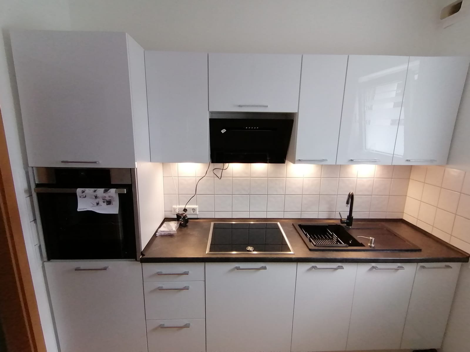 Biała kuchnia z czarnym blatem i sprzętami. Górne szafki lakierowane, podświetlane. Piekarnik w zabudowie. Nowoczesny design. Uwagę przykuwa czarna bateria.