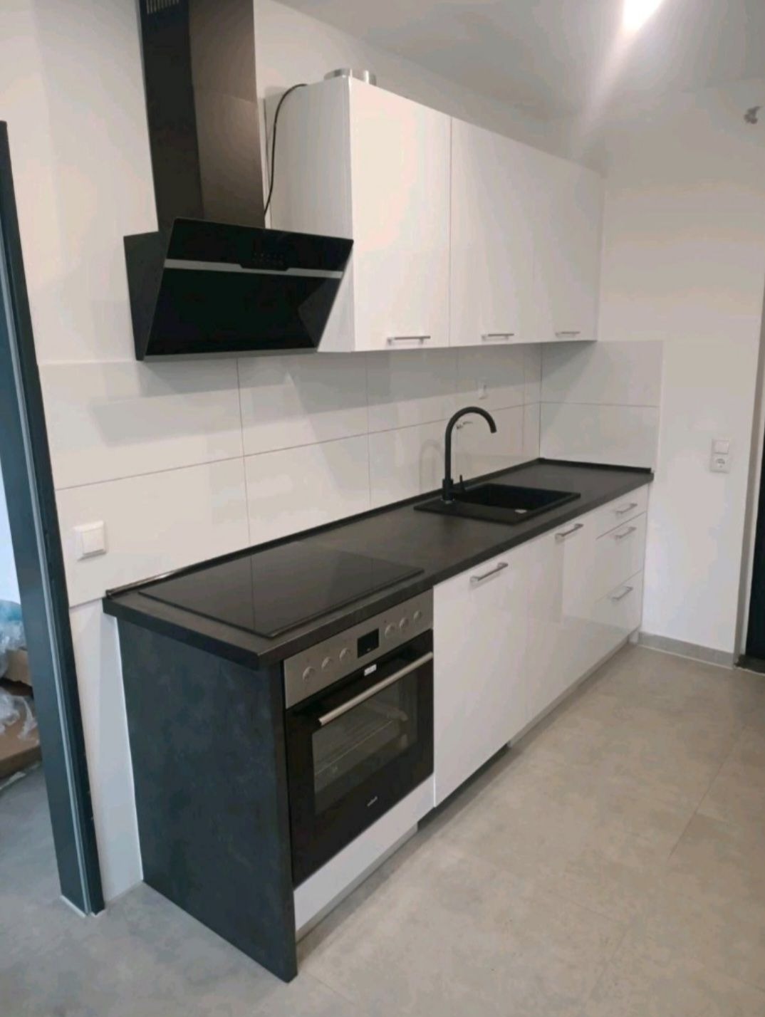 Nowoczesna kuchnia z białymi szafkami, czarnym blatem i okapem. Widoczny piekarnik i płyta indukcyjna. Minimalistyczny styl, jasne płytki na ścianie.