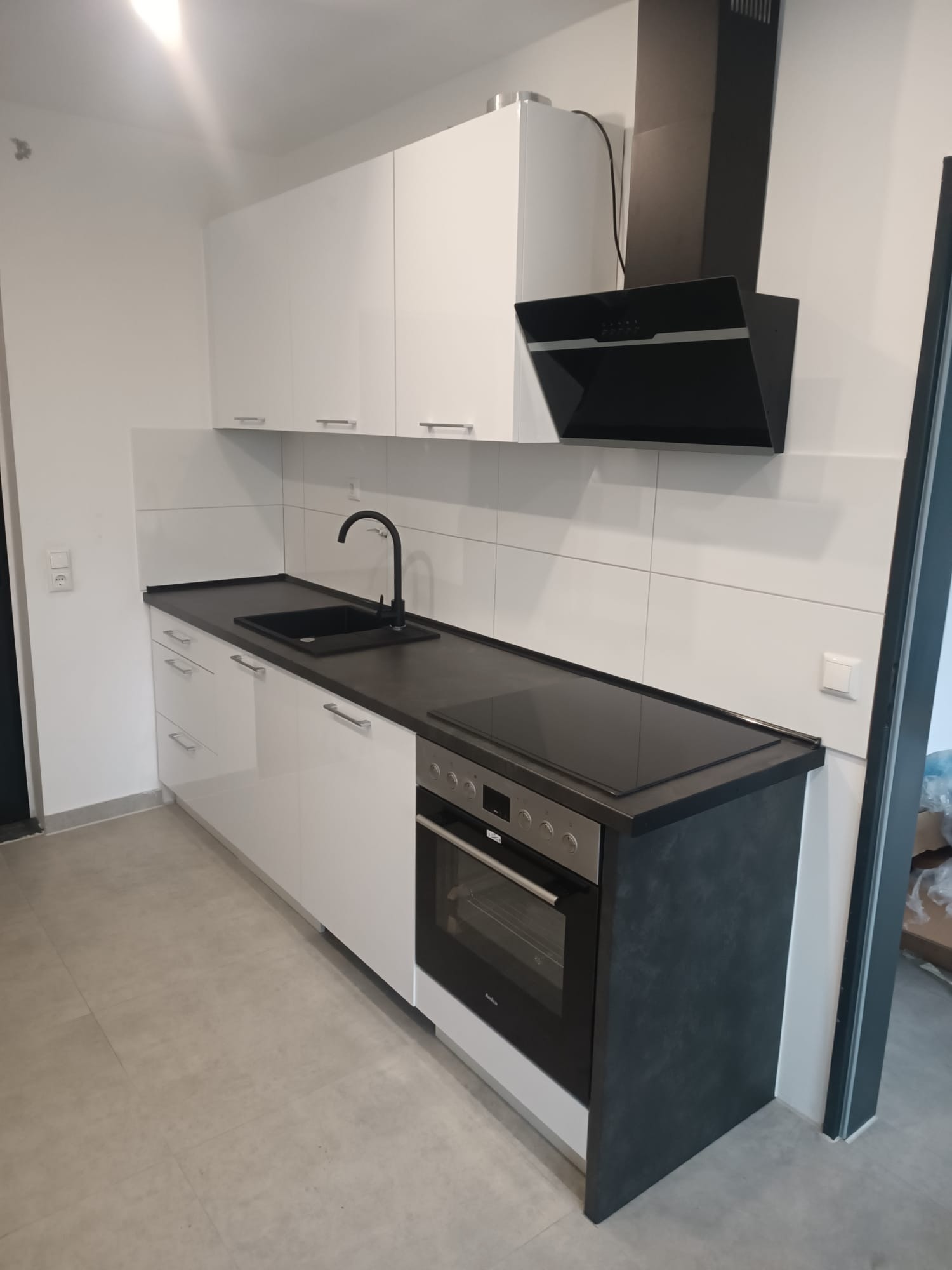 Jasna kuchnia z białymi szafkami, czarnym blatem i okapem. Minimalistyczny styl, proste uchwyty. Nowoczesne wykończenie.