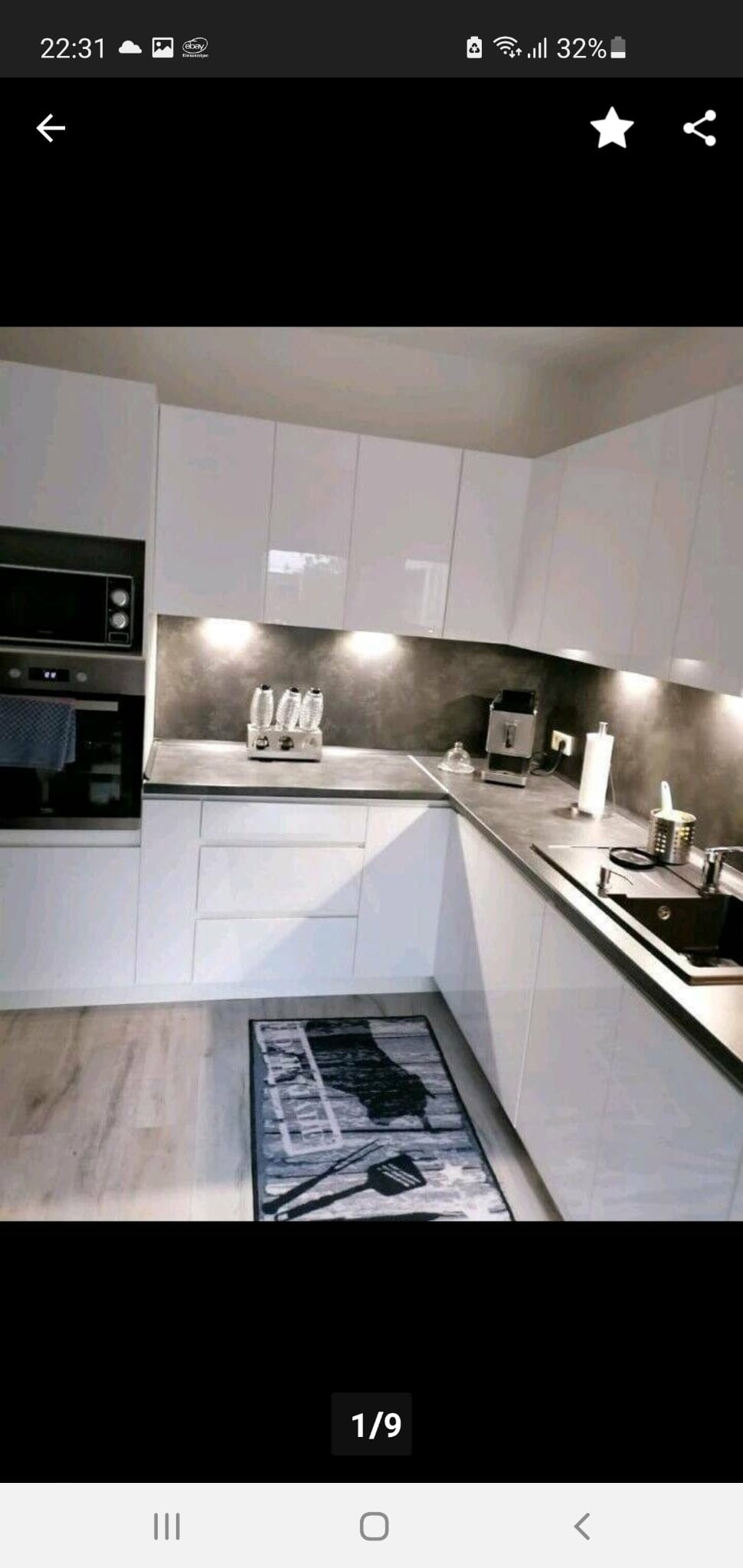 Nowoczesna, biała kuchnia z połyskiem, szary blat. Na blacie toster, ekspres do kawy. Na podłodze dywanik z motywem grilla. Oświetlenie punktowe pod szafkami.