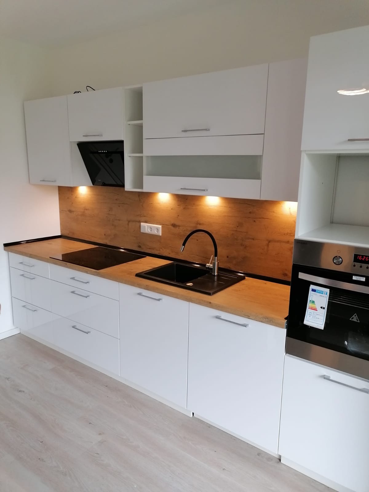 Nowoczesna, biała kuchnia z drewnianym blatem i czarnym zlewem. Nad blatem szafki i oświetlenie punktowe. Widoczny piekarnik ze srebrnym wykończeniem.