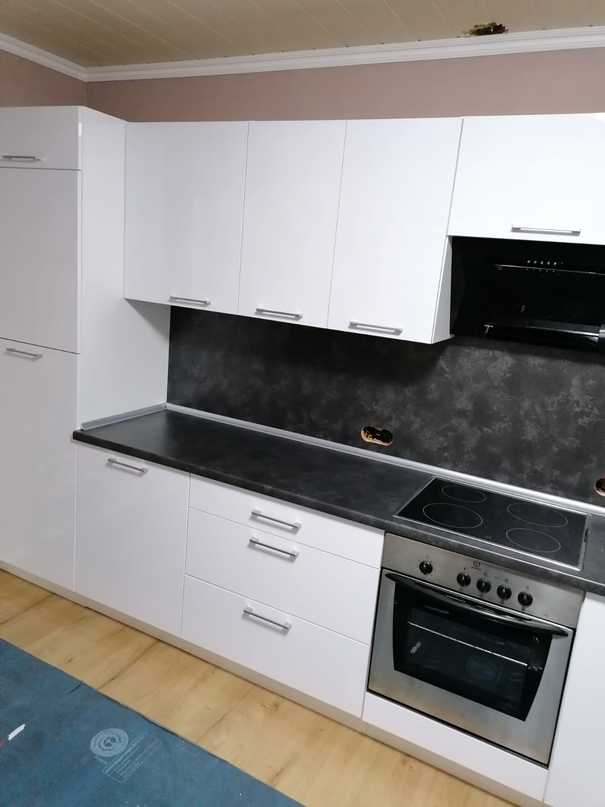 Biała, lakierowana kuchnia z czarnym blatem i płytą indukcyjną. Górne szafki wiszące, dolne z szufladami. Tło stanowi ściana w odcieniach szarości.