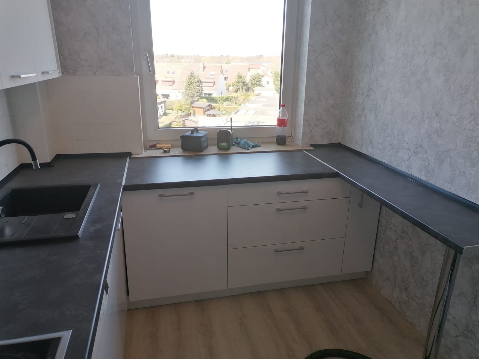 Jasna kuchnia z szarym blatem i widokiem z okna na zabudowę jednorodzinną. Na blacie narzędzia i akcesoria, co sugeruje montaż.