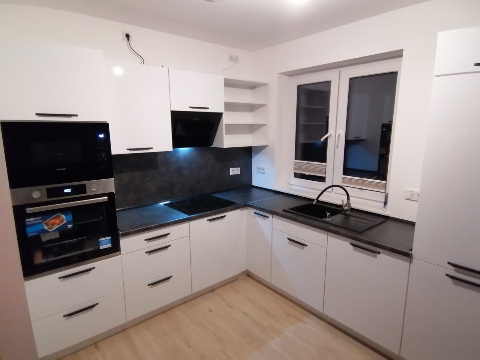 Nowoczesna, jasna kuchnia z białymi frontami i czarnym blatem. Widoczny piekarnik z wyświetlaczem i płyta indukcyjna. Uwagę zwracają minimalistyczne uchwyty.