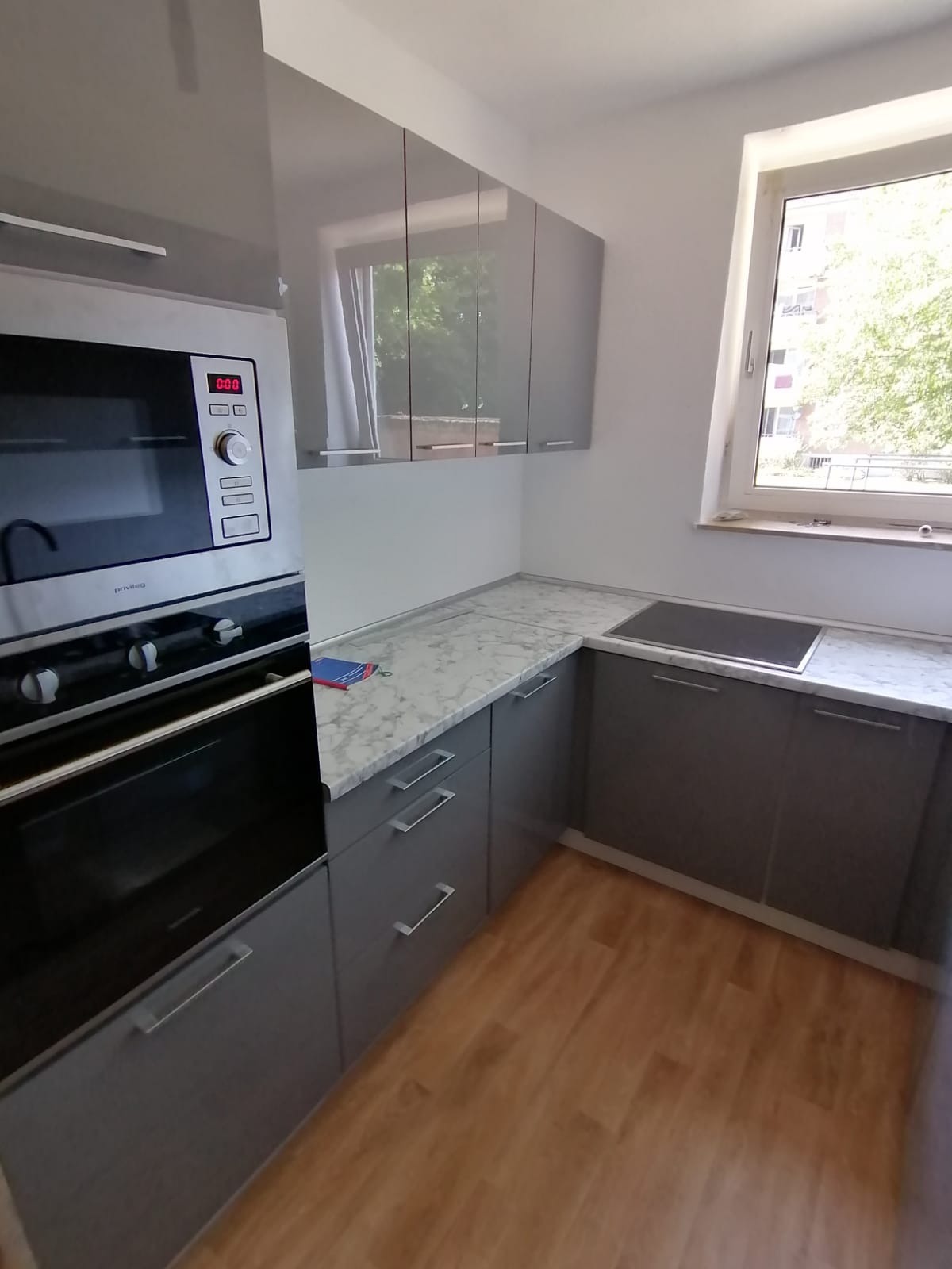 Szara kuchnia z marmurowym blatem, zabudowana piekarnikiem i szafkami wiszącymi, z widokiem na zieleń za oknem. Nowoczesne wykończenie, drewniana podłoga.