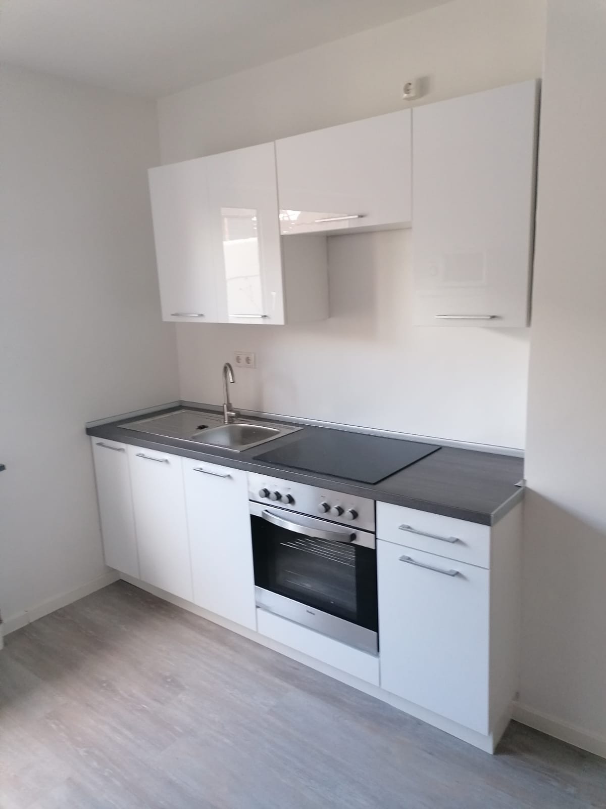 Nowoczesna, biała kuchnia z szarym blatem, z zabudowaną płytą grzewczą i piekarnikiem. Minimalistyczny design, widok z lekko podwyższonej perspektywy.