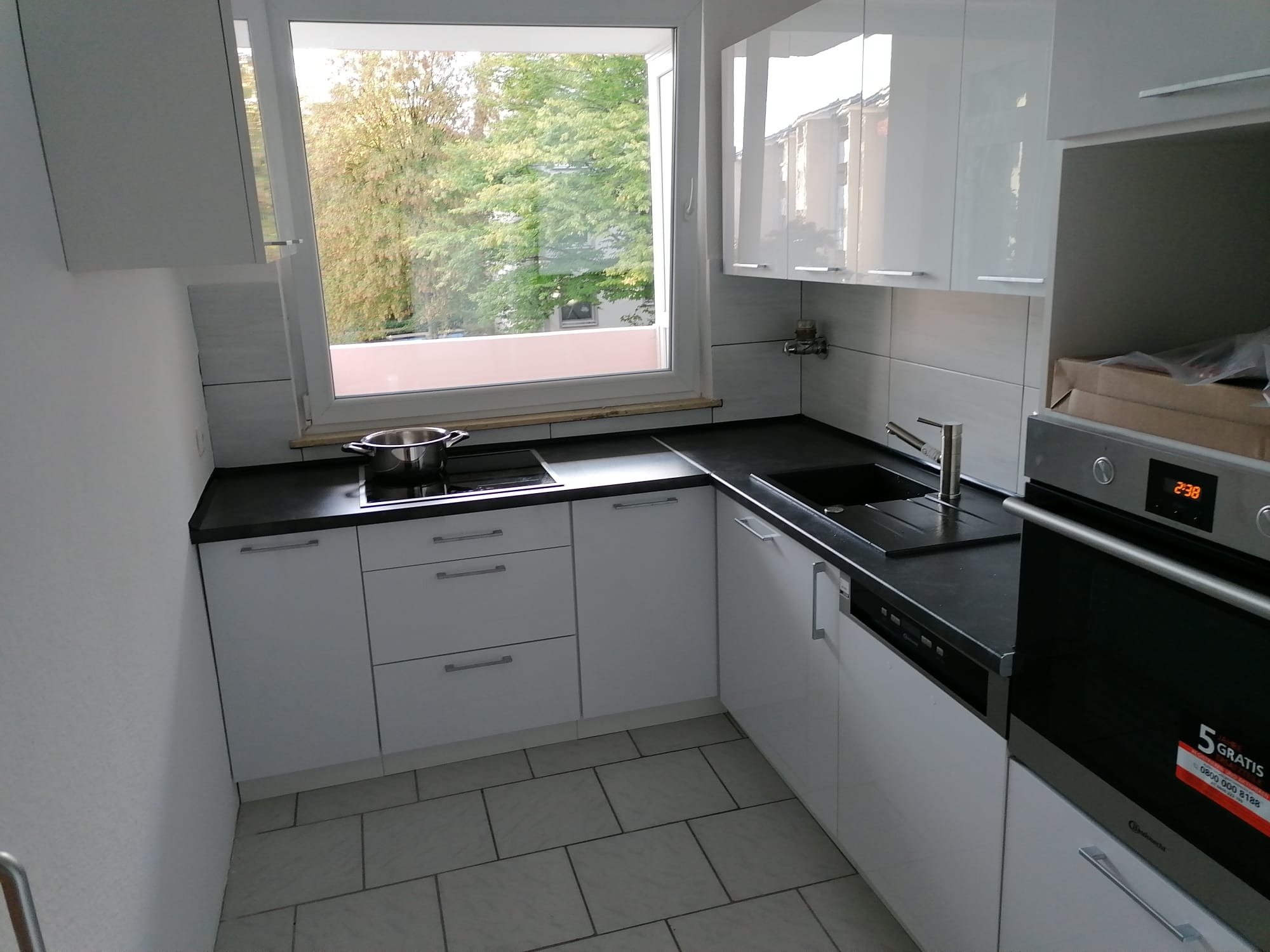 Nowoczesna, jasna kuchnia z białymi szafkami i czarnym blatem. Widoczne okno z widokiem na zieleń i fragment piekarnika z wyświetlaczem. Minimalistyczny design.