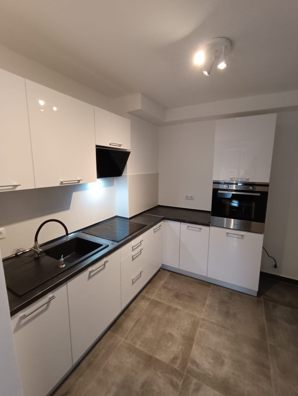 Biała, nowoczesna kuchnia z czarnym blatem i sprzętem AGD w zabudowie. Minimalistyczny design, oszczędne oświetlenie punktowe.