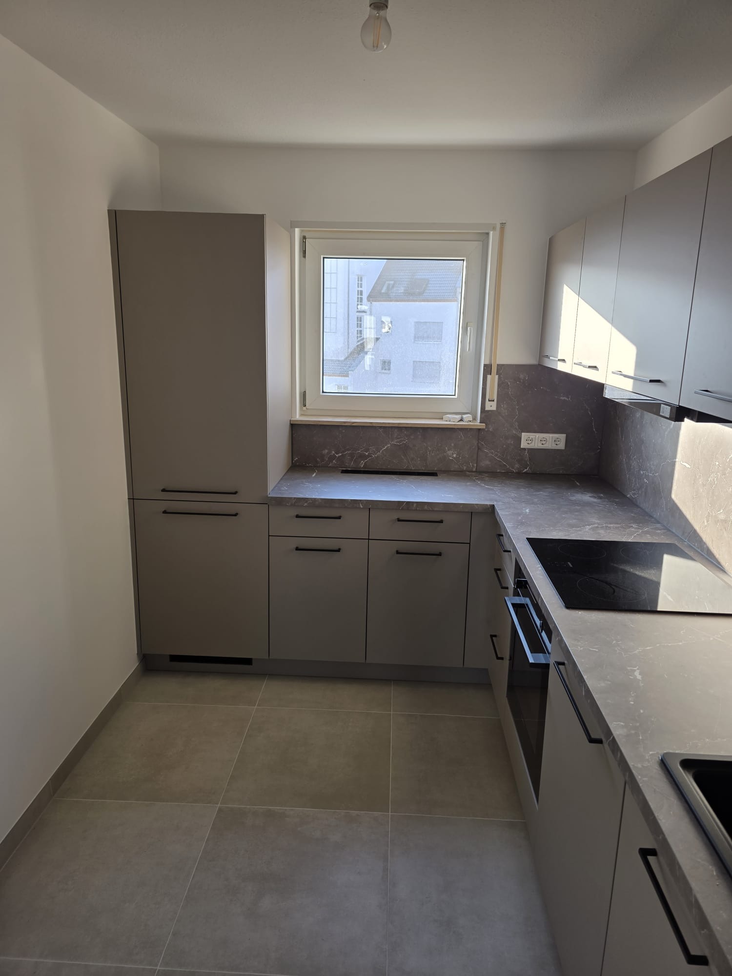 Szara, nowoczesna kuchnia z czarnymi uchwytami i marmurowym blatem. Widok na okno z widokiem na budynek. Minimalistyczny design, oświetlenie sufitowe.