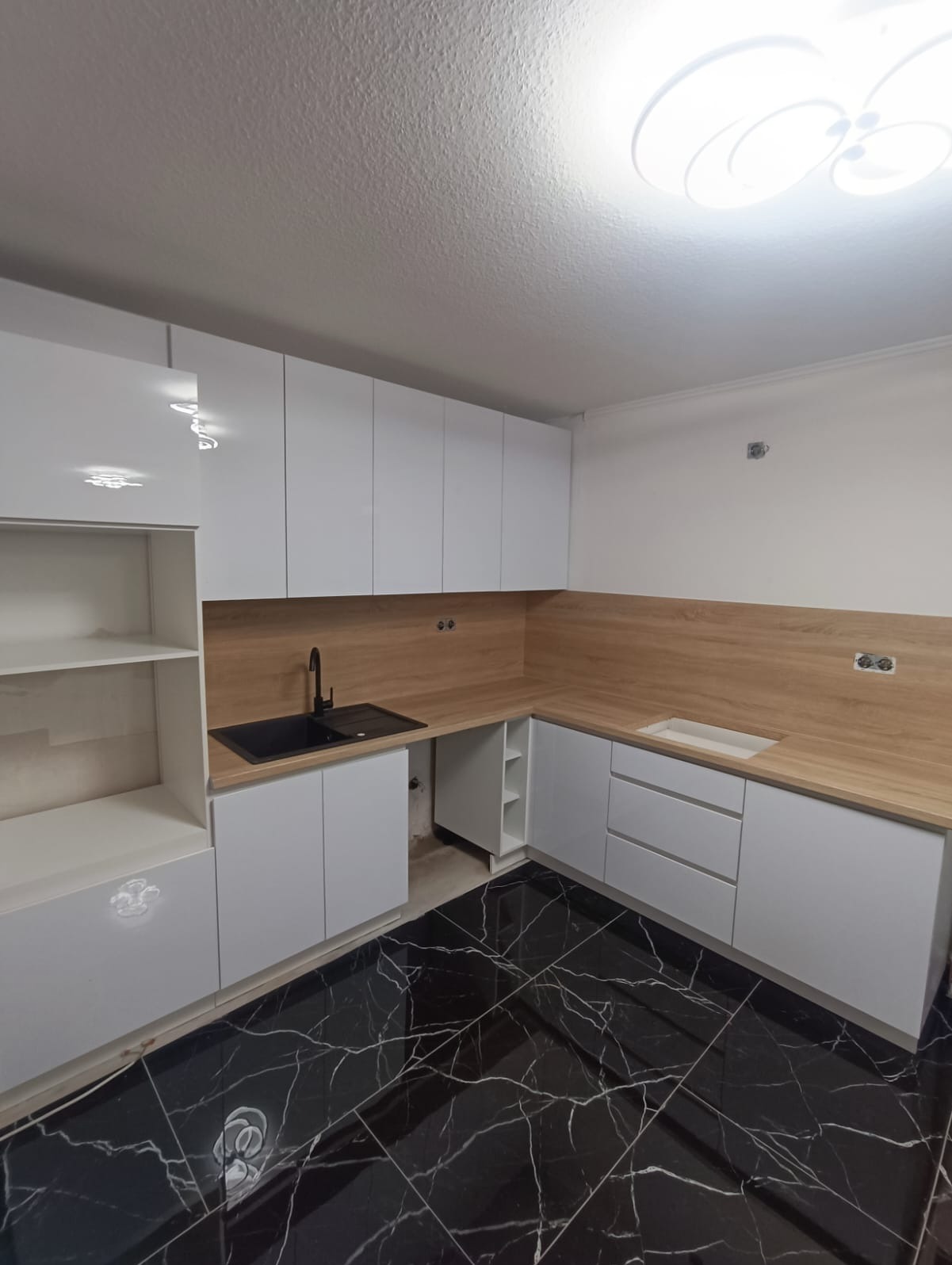 Nowoczesna kuchnia z białymi szafkami i drewnianym blatem, czarny zlew i marmurowa podłoga. Minimalistyczny design, oświetlenie sufitowe.