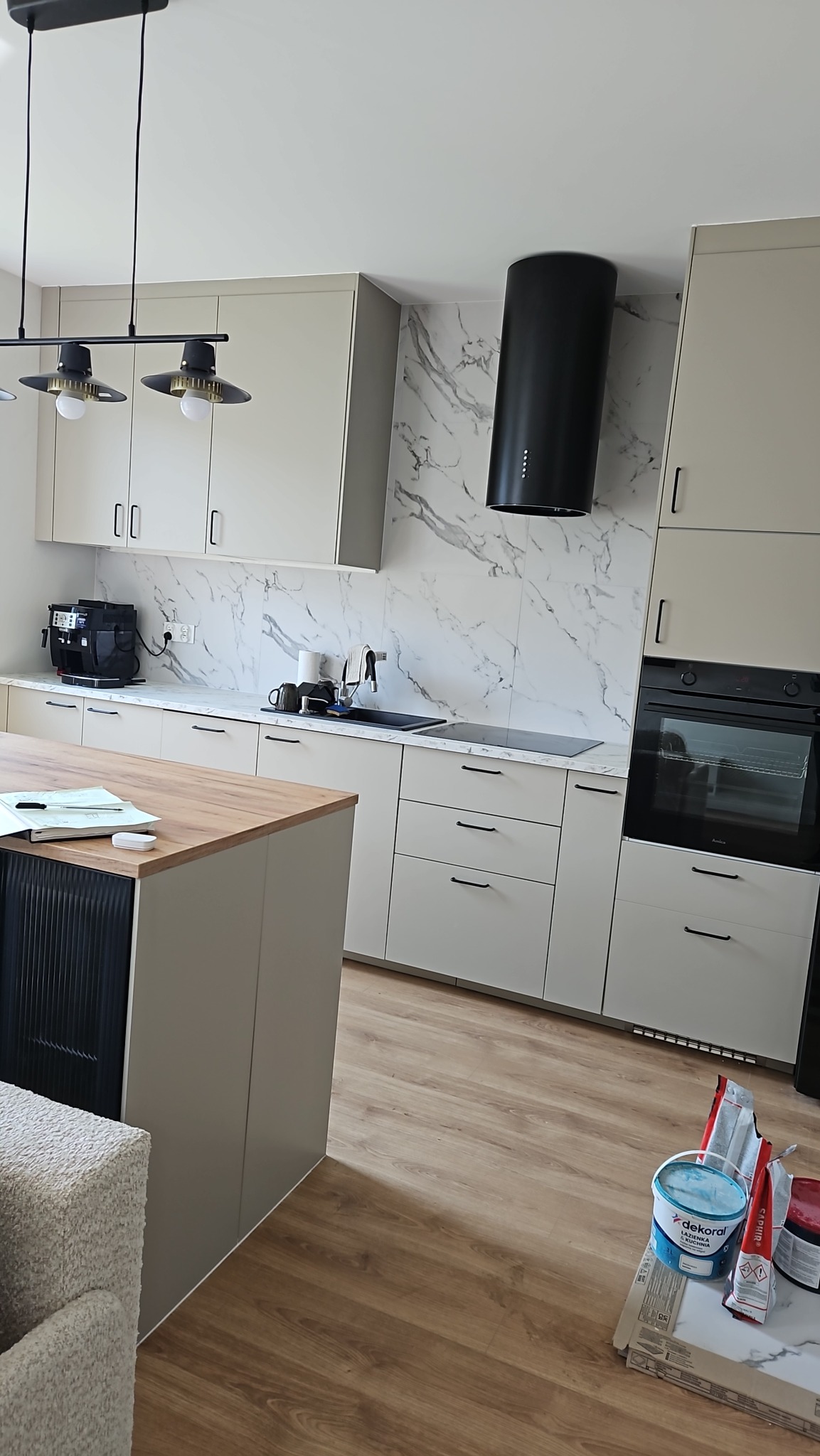 Jasna kuchnia z wyspą i marmurowym backsplashiem. Czarny okap cylindryczny i minimalistyczne lampy wiszące. Na podłodze panele, widoczne farby i płytki.