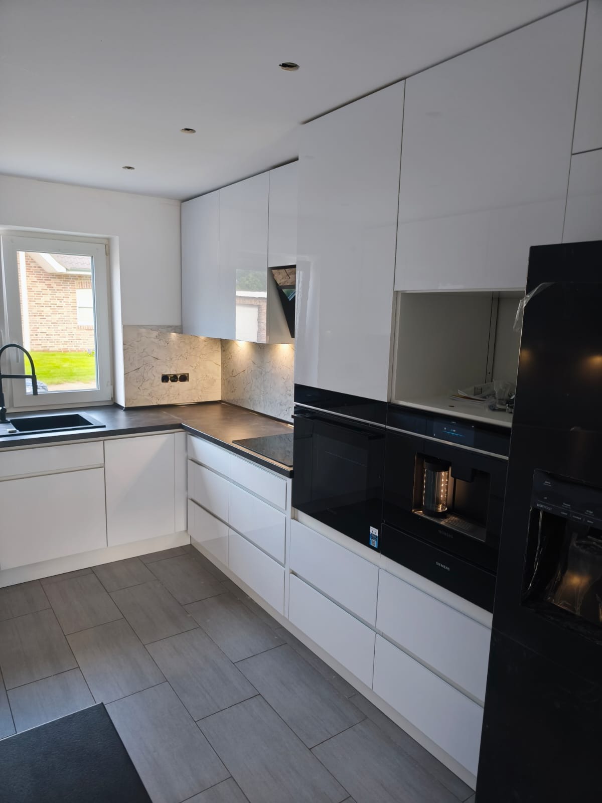 Biała, nowoczesna kuchnia z błyszczącymi frontami, szarym blatem i marmurowym panelem nad nim. W zabudowie piekarnik i ekspres do kawy. Widok na okno i fragment podłogi.