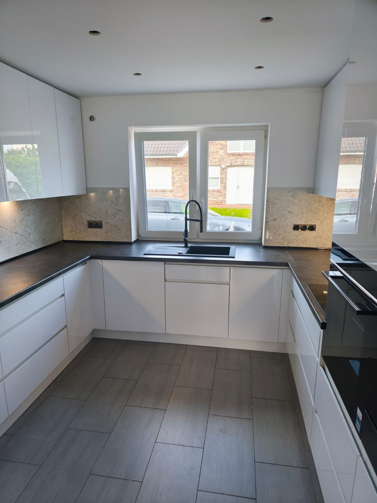 Biała, nowoczesna kuchnia z czarnym blatem i zlewozmywakiem, marmurowym panelem nad blatem, widok na okno z zabudową domów w tle.