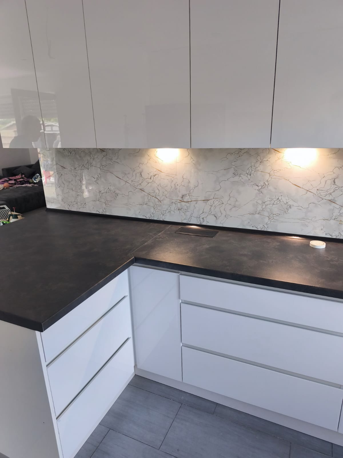 Biała, lakierowana zabudowa kuchenna z ciemnym blatem i marmurowym panelem nad blatem, oświetlonym punktowo. Widoczny fragment salonu w tle.