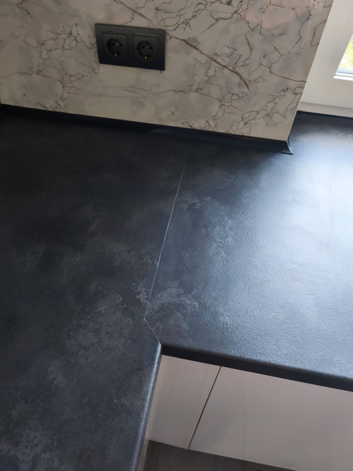 Czarny blat kuchenny z widocznym łączeniem, marmurowa ściana nad blatem i gniazdka elektryczne. Fragment białych szafek kuchennych w dolnej części kadru.
